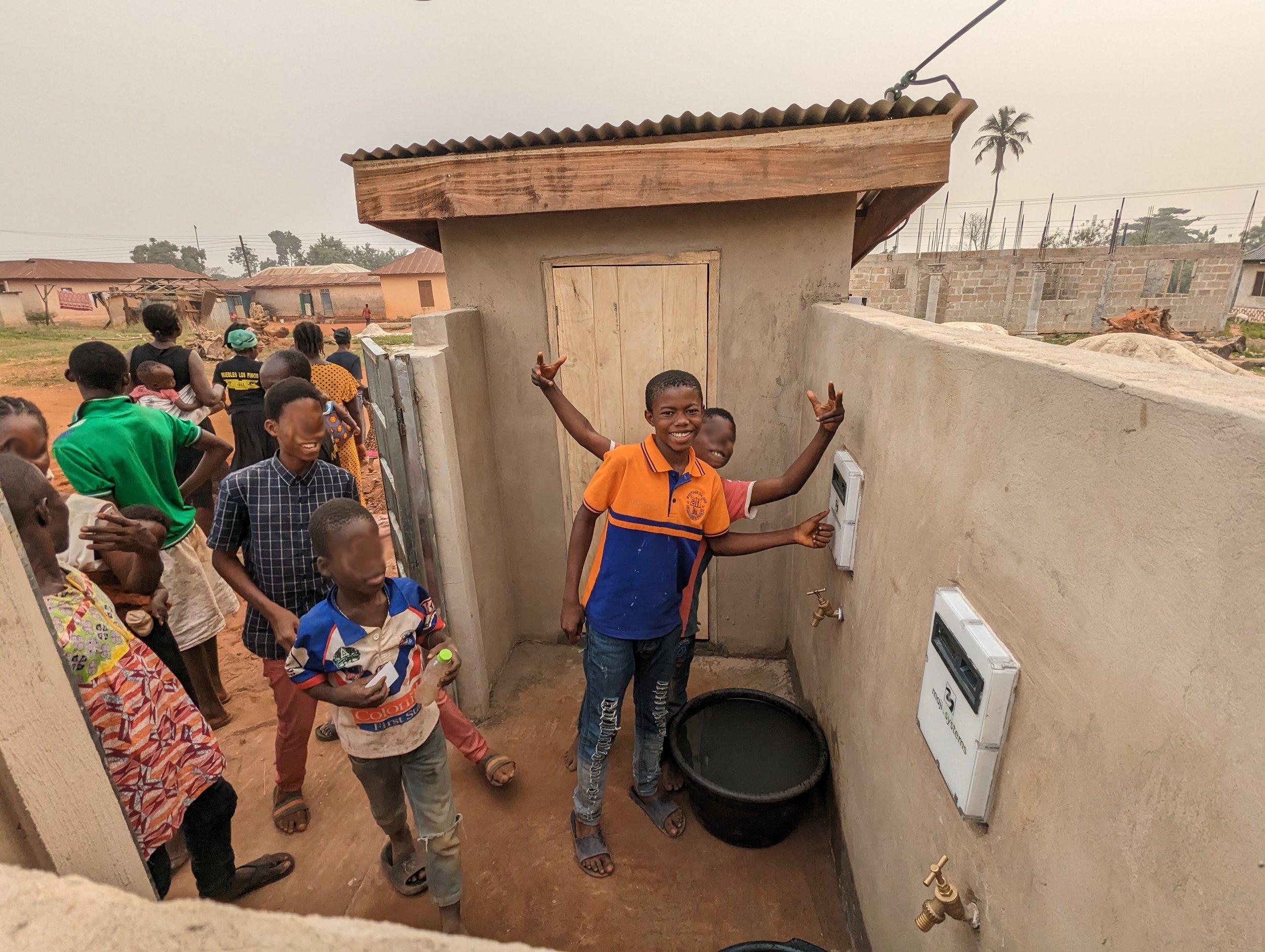 Children at a village standpipe in the Ashanti Region, Ghana, one smiling beside two wall-mounted Maji Community Dispensers