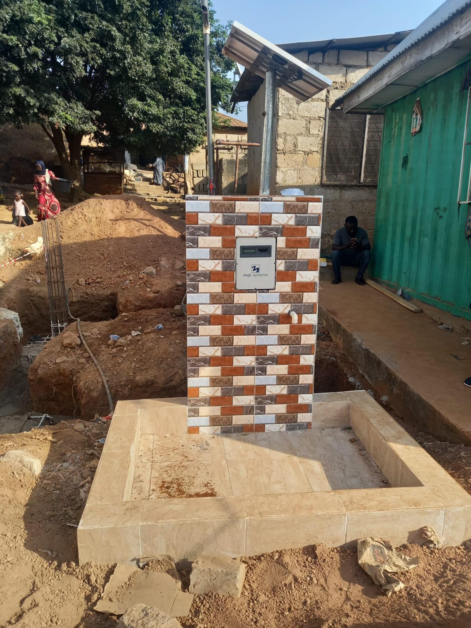 A solar-powered Maji Community Dispenser set into a tiled standpipe in a Kumasi neighbourhood, with a woman and child walking past