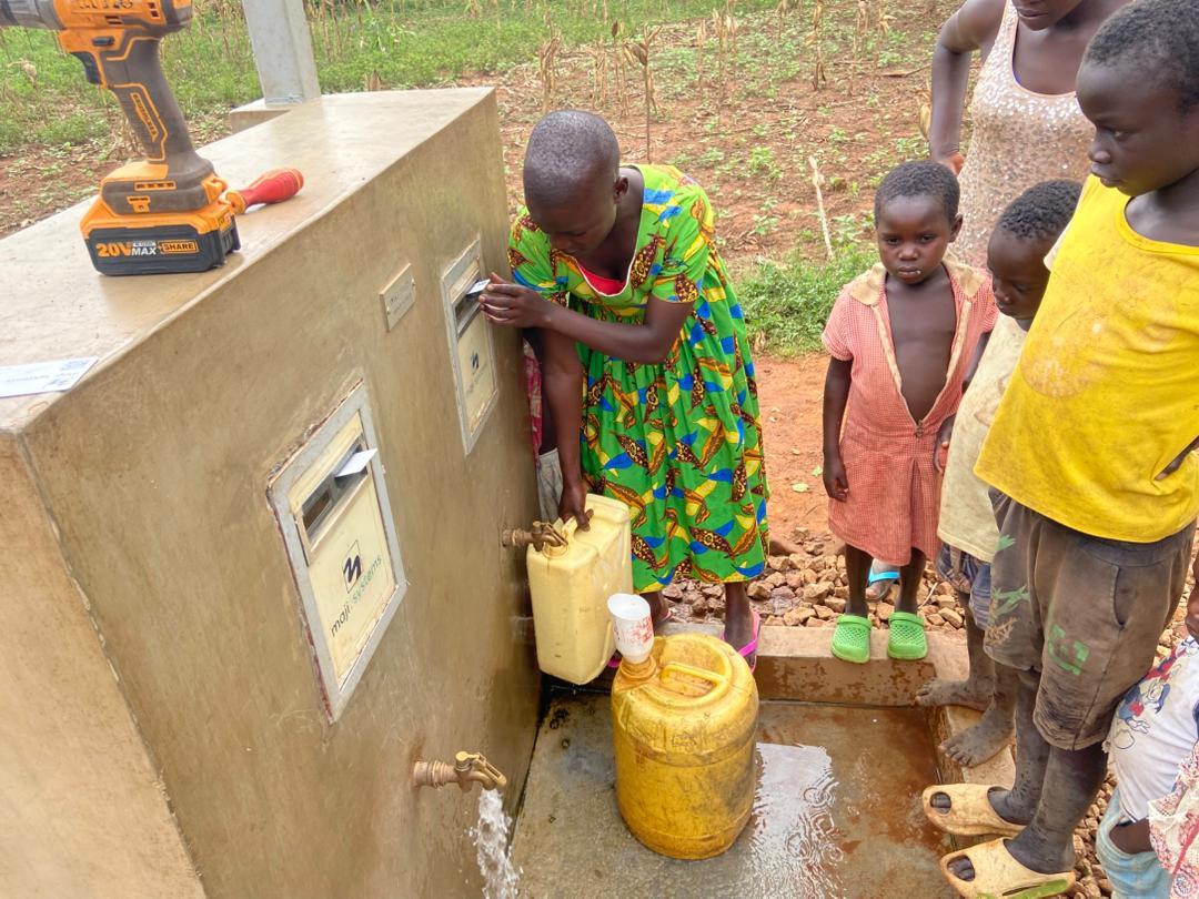 A woman filling a yellow jerrycan at a Maji Community Dispenser while children watch