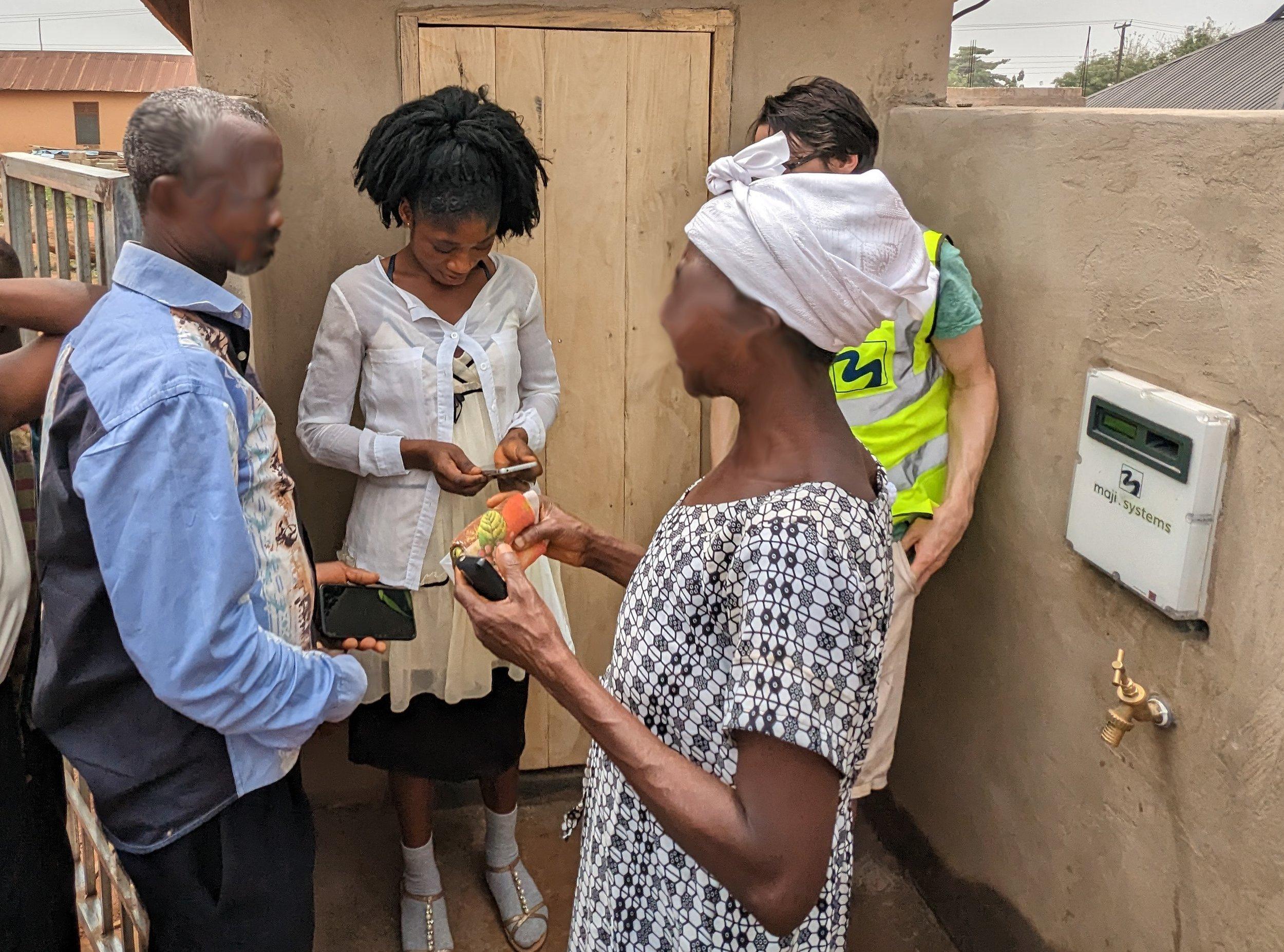 A reseller topping up a customer’s WaterCard on her phone beside a wall-mounted Maji dispenser